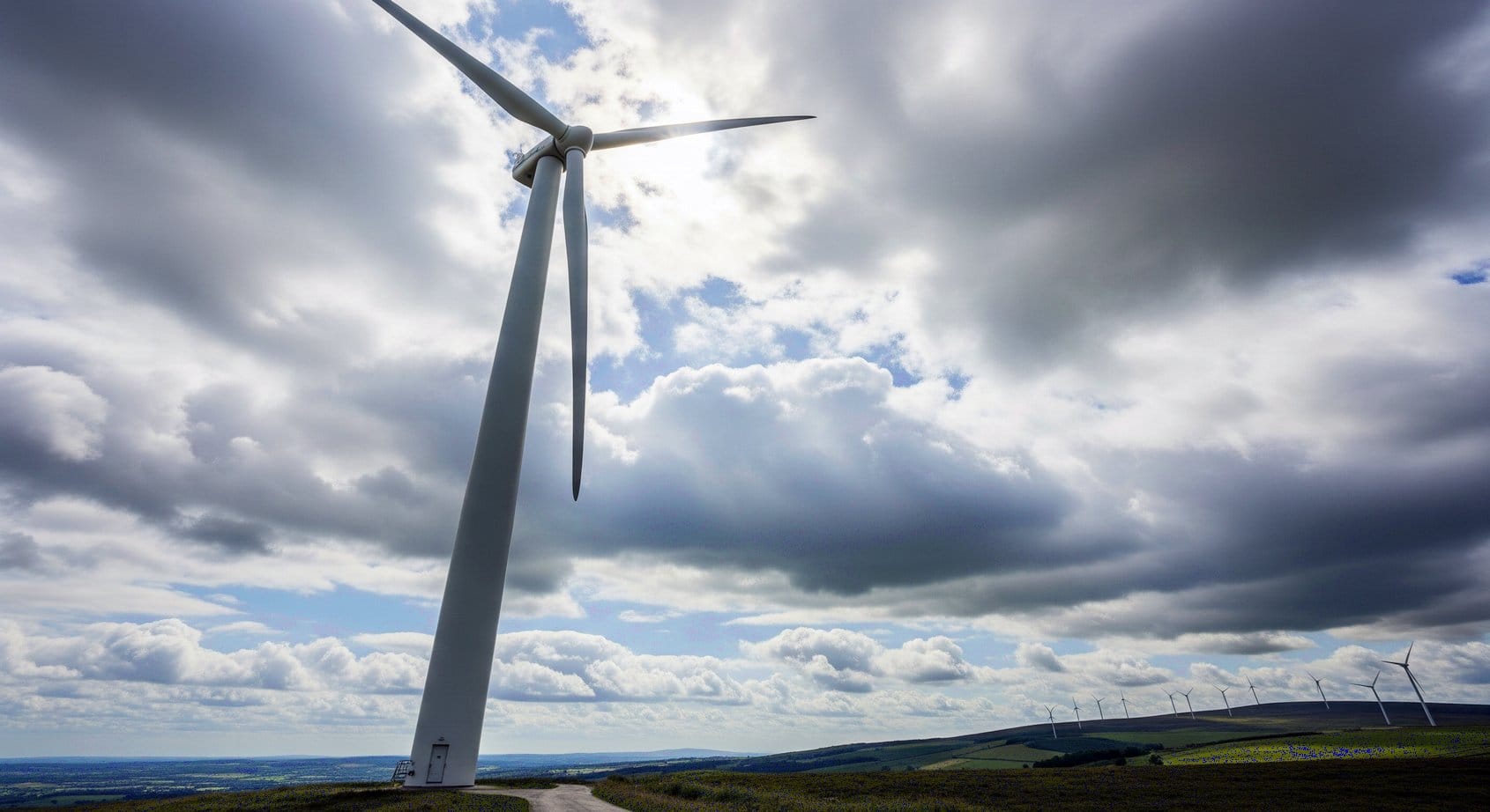 A tall, modern wind turbine with three white blades stands against a cloudy sky, generating renewable energy